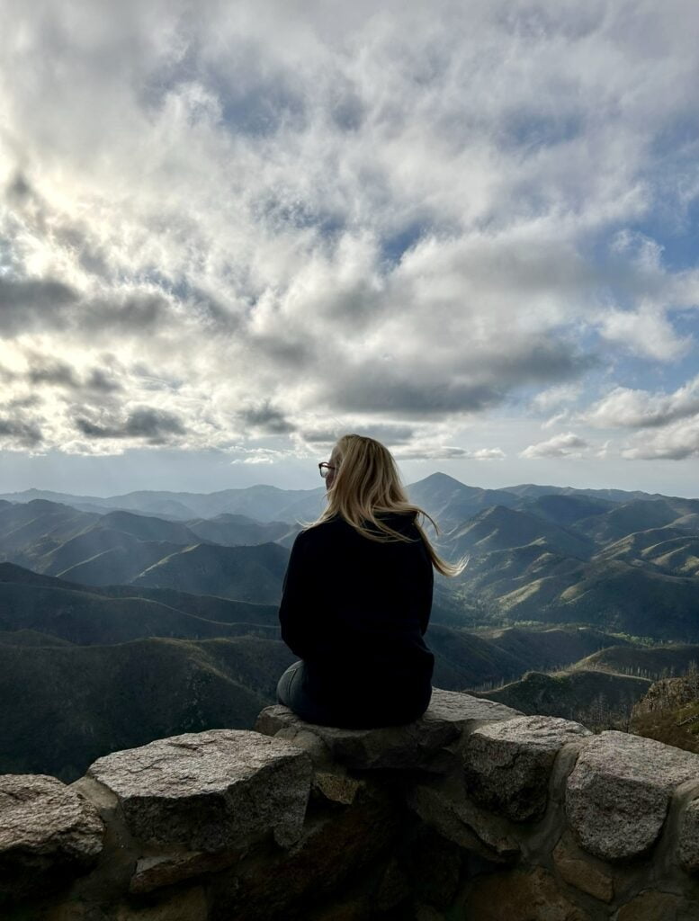A solo camping woman sits on a rock overlooking the mountains.
